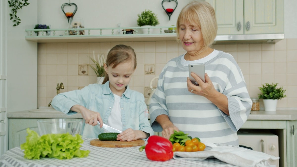 Grandmother with smartphone is teaching granddaughter to cook salad at home, cute little girl is cutting cucumber using knife and chopping board.