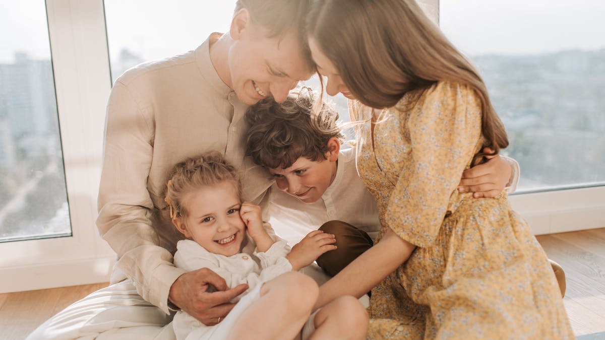 Cheerful family of four cuddling and smiling together in a bright, sunlit living room setting.